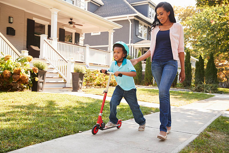 Mother Walks With Son As He Rides Scooter Along Sidewalk